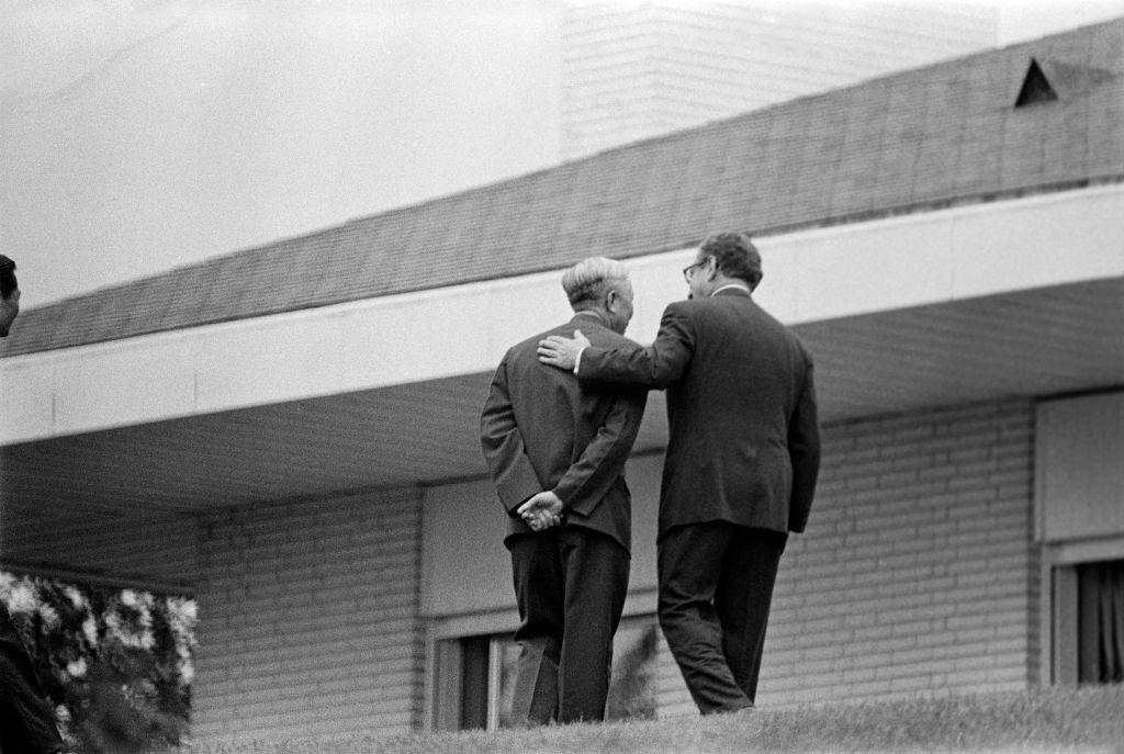 Henry Kissinger pone una mano en la espalda de Le Duc Tho mientras caminan hacia una casa