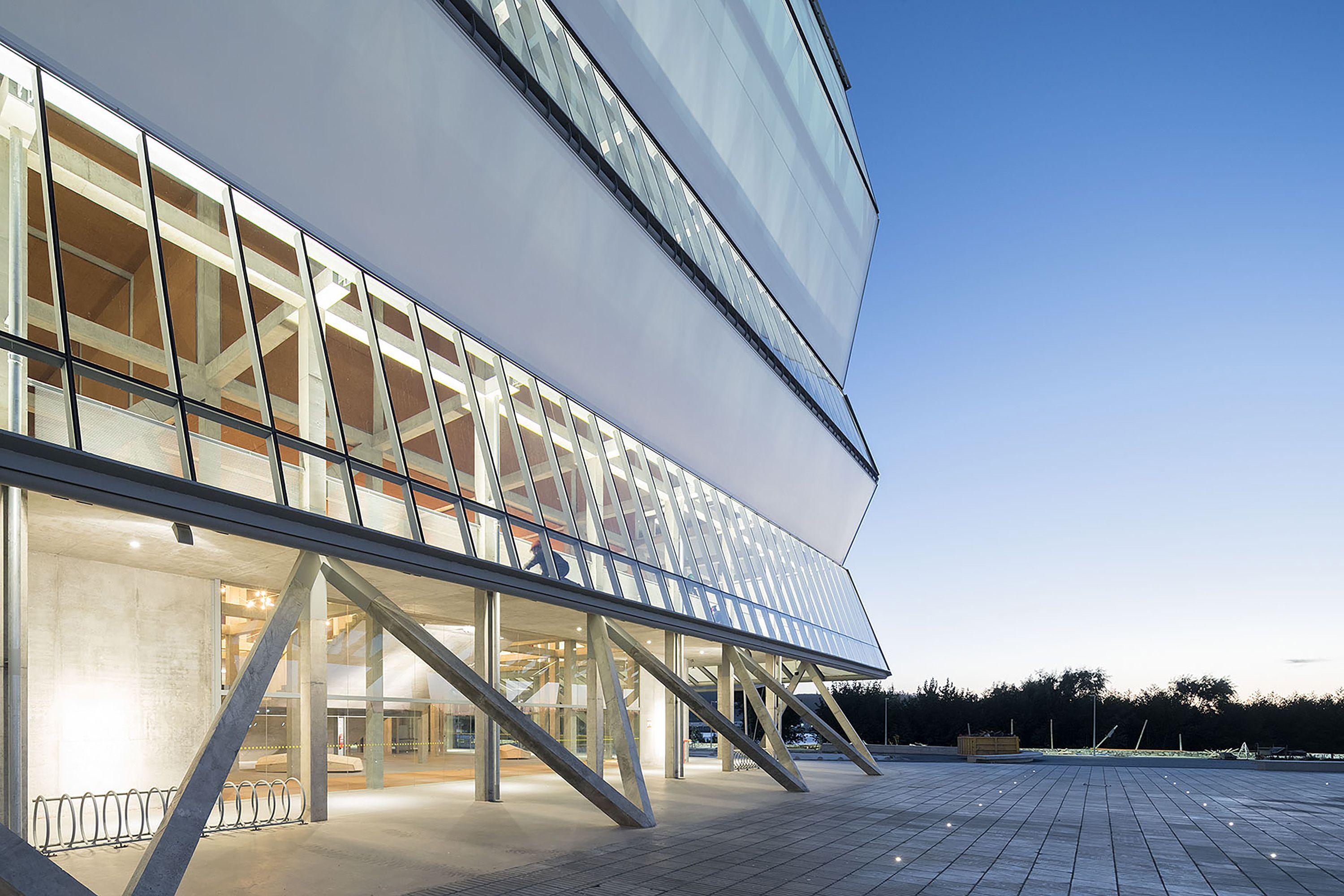 Fachada inclinada de un edificio moderno con estructura metálica expuesta y ventanales amplios al atardecer.