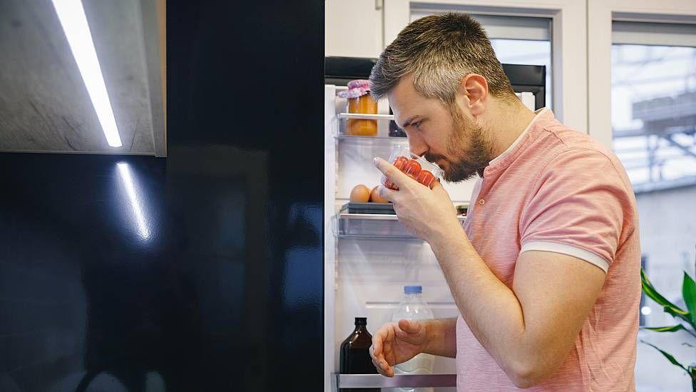Hombre frente a su refrigerador oliendo un recipiente con alimentos