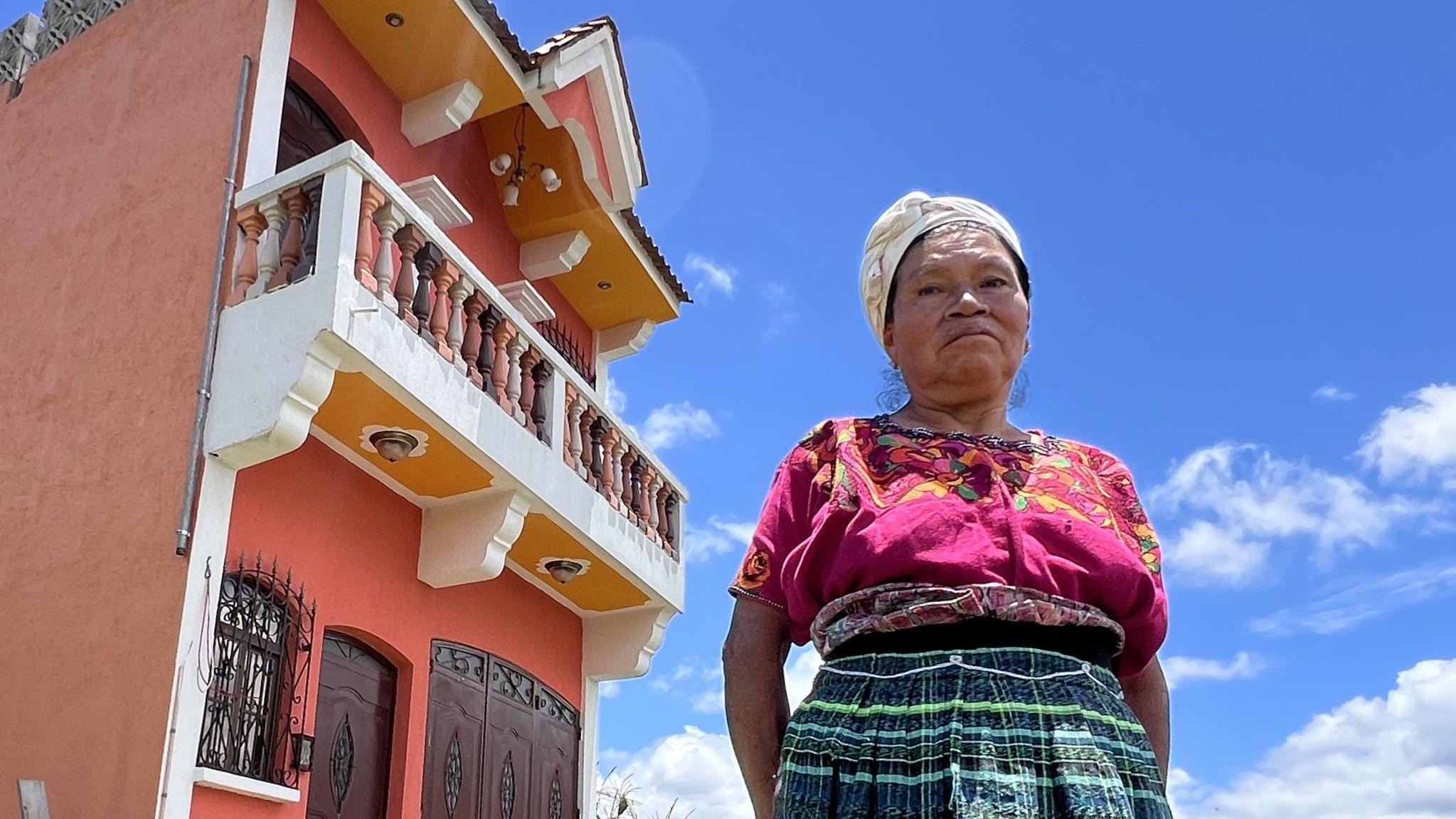 Mujer vestida con ropa de colores, con el pelo recogido y mirando a cámara desde lo alto. Detrás, se ve la fachada de su casa de remesas, con un balcón grande y colores variados.
