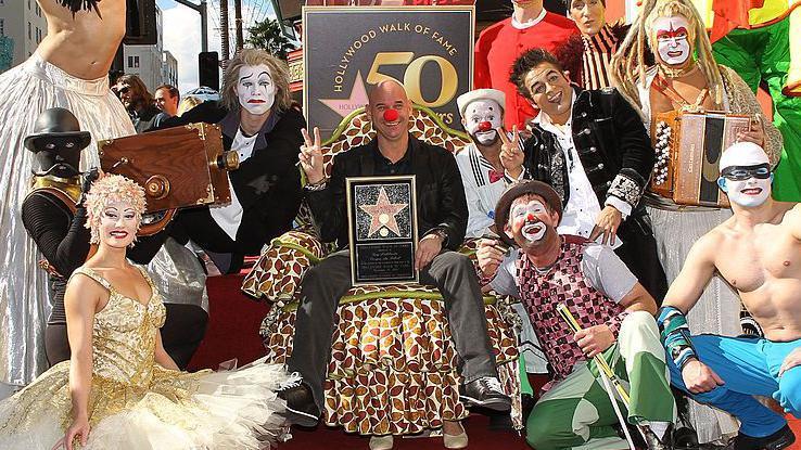El fundador del Cirque du Soleil, Guy Laliberté, recibe su estrella en el Paseo de la Fama de Hollywood. Sentado y sonriente, sostiene la estrella enmarcada, con una nariz roja de payaso y haciendo la señal de victoria,