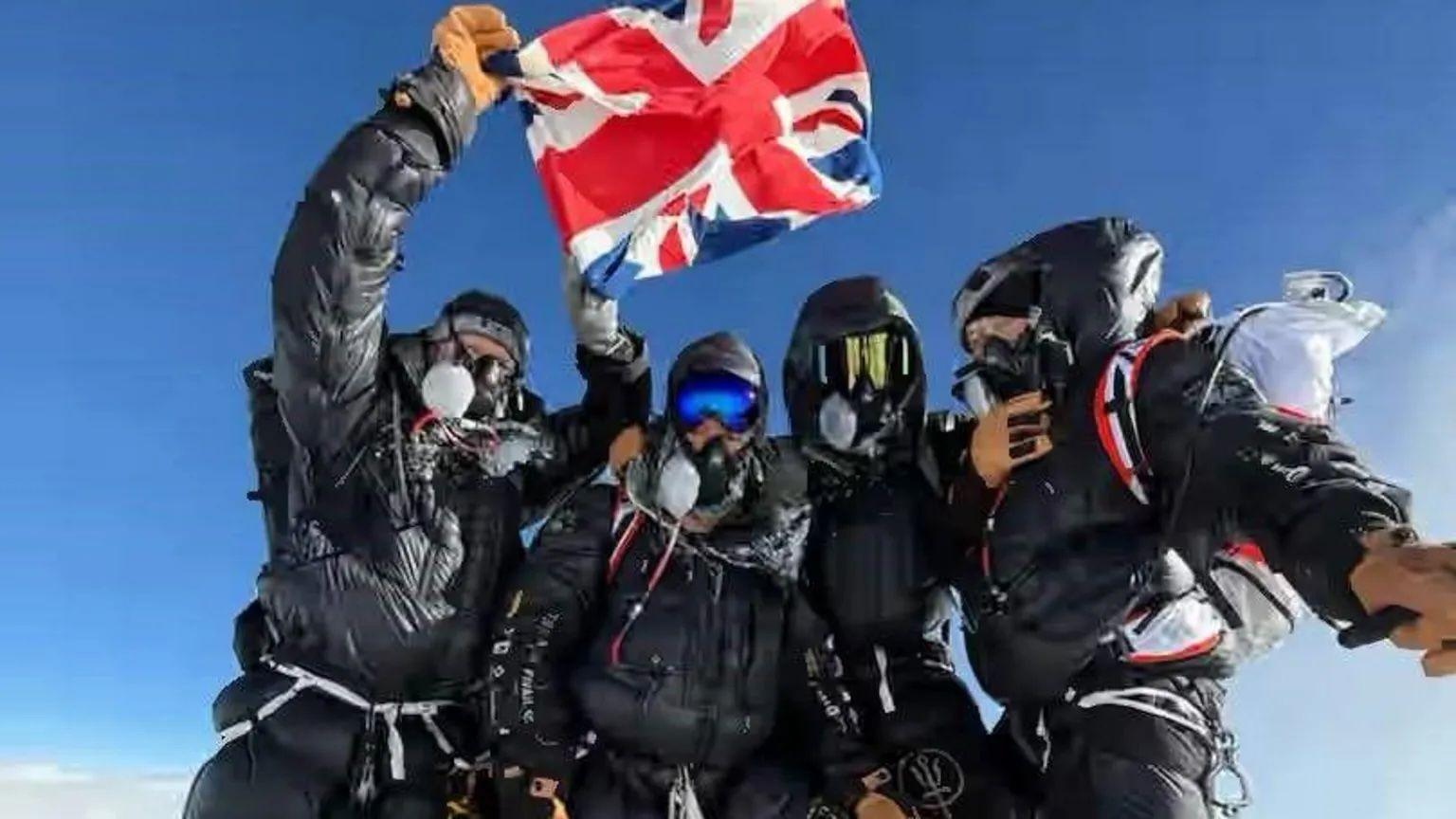 Cuatro escaladores juntos, vestidos con equipo de escalada y máscaras, parados en una cima mientras uno sostiene una bandera del Reino Unido