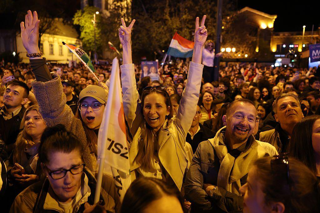 Un grupo de personas celebran en la noche de Budapest el triunfo de Peter Magyar. Llevan banderas del partido Tisza y al fondo ondea una bandera húngara. 