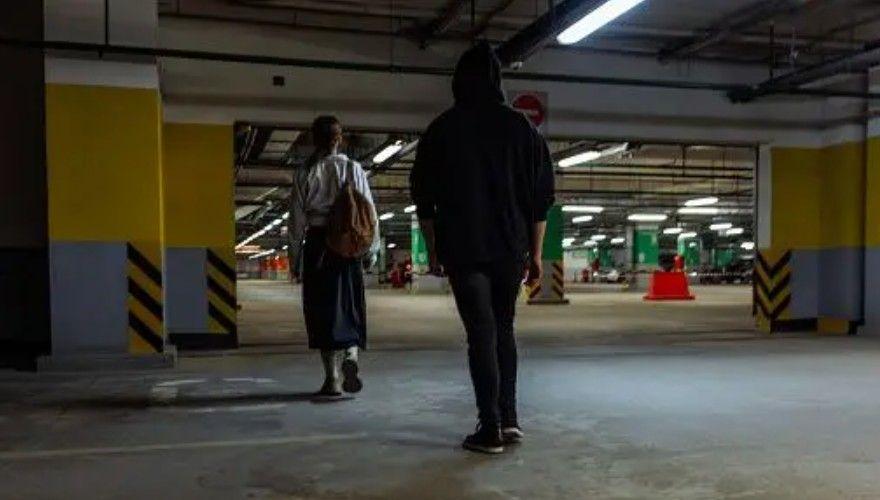 Un hombre con la capucha subida sigue a una mujer por un aparcamiento subterráneo tenuemente iluminado.