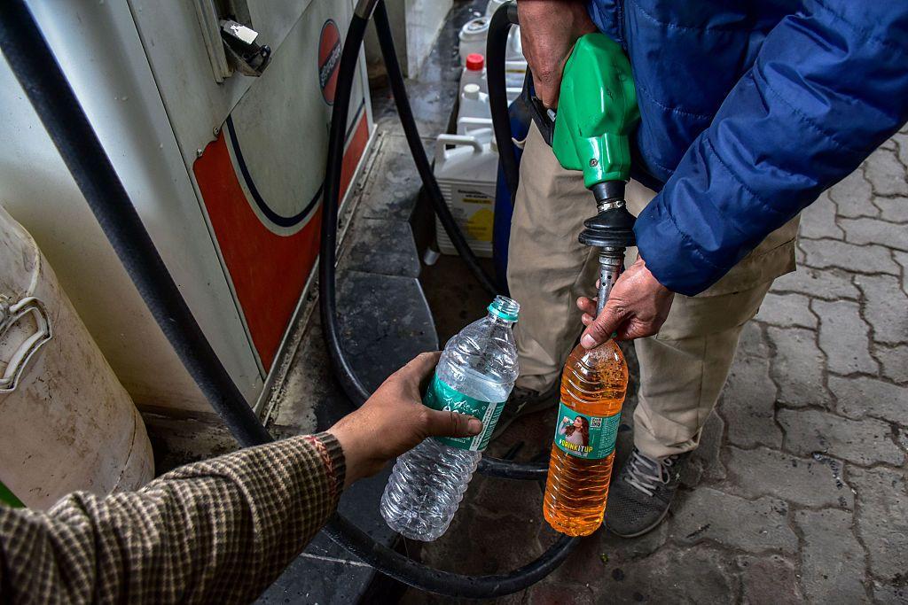 Um frentista vestindo uma jaqueta puffer azul enche uma garrafa pl&aacute;stica com gasolina em um posto de combust&iacute;vel, enquanto a outra m&atilde;o segura uma garrafa vazia.