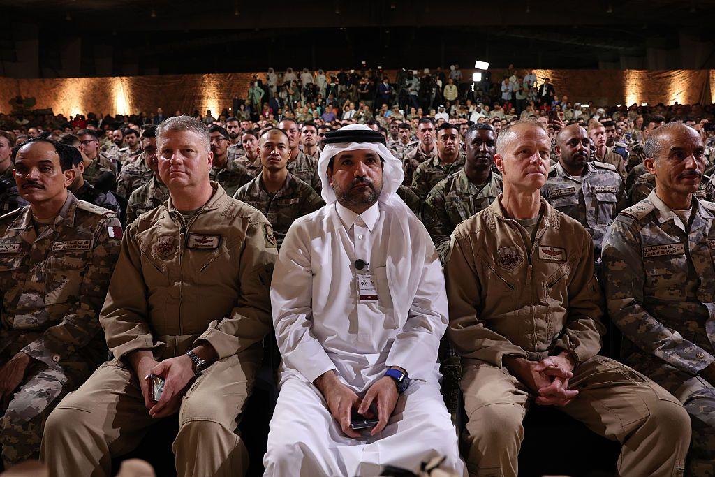 لماذا اختارت إيران القاعدة الأمريكية في قطر لتوجيه رسالتها لواشنطن؟ 2 عسكريون أمريكيون وقطريون يستعمون إلى الرئيس الأمريكي دونالد ترامب وهو يتحدث على خشبة المسرح خلال جولة في قاعدة العديد الجوية في 15 مايو 2025، في العاصمة القطرية الدوحة.