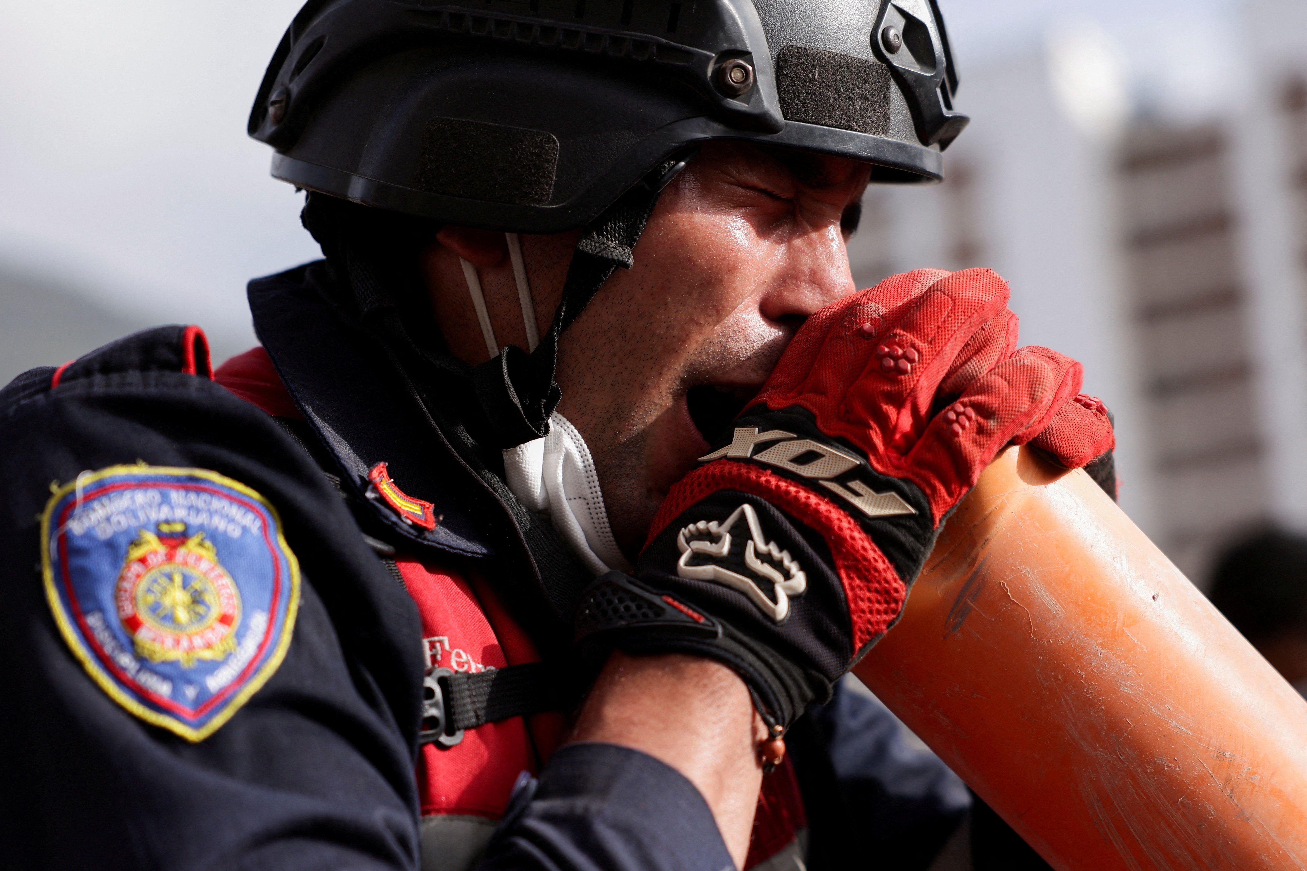 Un rescatista con casco, uniforme, guantes rojos y un tapabocas mal puesto, que lleva debajo de la barbilla, grita por un tubo color naranja de PVC. 