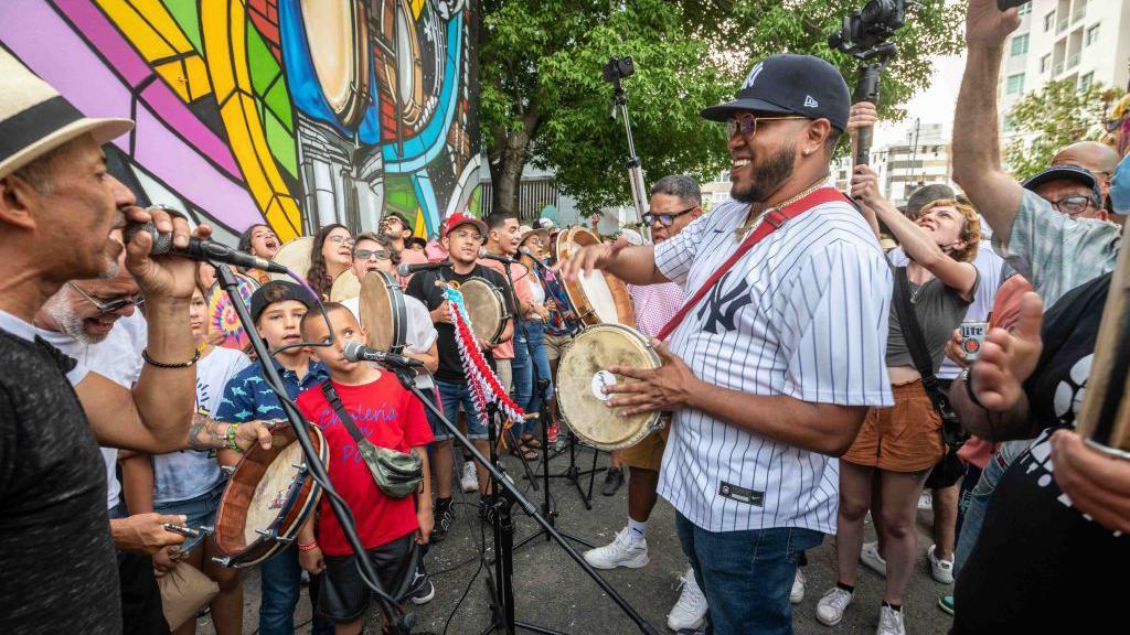 varias personas tocan panderos de bomba y cantan rodeados de niños en puerto rico. 