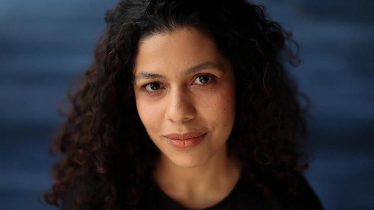 Arianna de Souza-García mira a la cámara. Es una mujer de cabello rizado y oscuro, con camiseta negra, situada frente a un fondo azul oscuro.