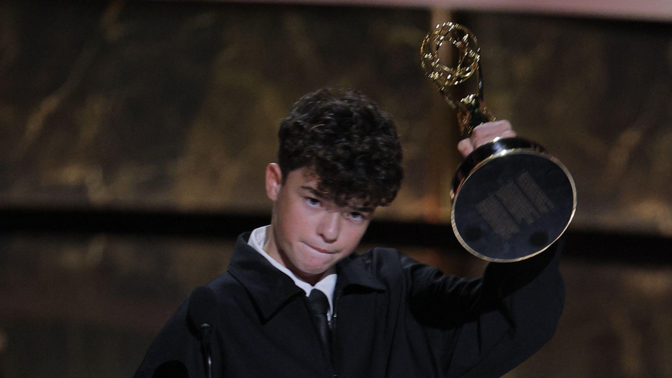 Owen Cooper vestido con chaqueta negra y camisa blanca, levanta con su mano izquierda el trofeo por ganar el Emmy.