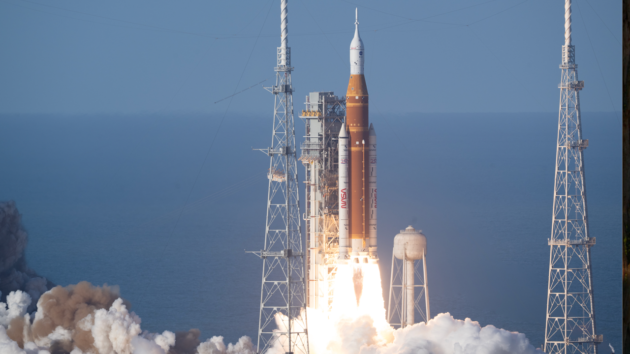 一枚高聳的白色與橘色火箭正從海岸的發射台升空。它的底部噴出火焰和耀眼的白色噴流，形成一條寬闊、發光的尾焰，將地面遮在濃厚的煙霧雲層之中。這枚火箭纖細得像鉛筆，筆直地指向清澈的藍天；在它身後，海面隱約可見。