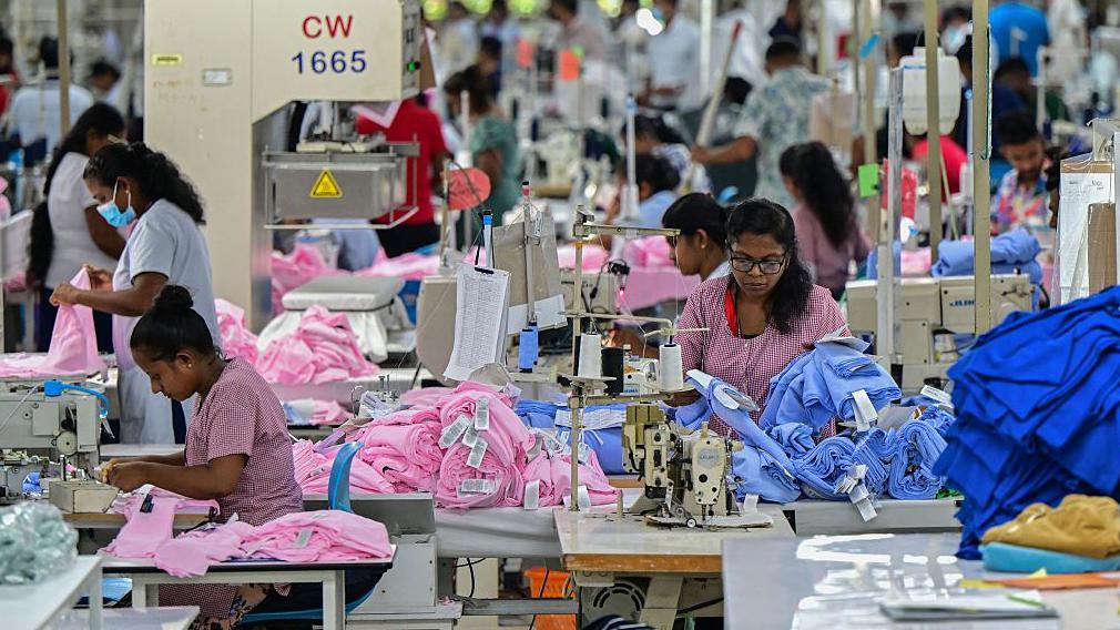 Empregados costurando roupas dentro de uma fábrica na zona franca de Katunayake, no Sri Lanka, com pilhas de tecidos azul e rosa ao lado das máquinas de costura