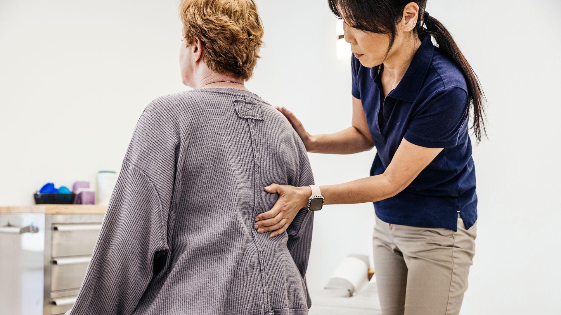 Una profesional de la salud tiene la mano en la espalda de una mujer mientras la examina, sentada en una sala de la clínica.