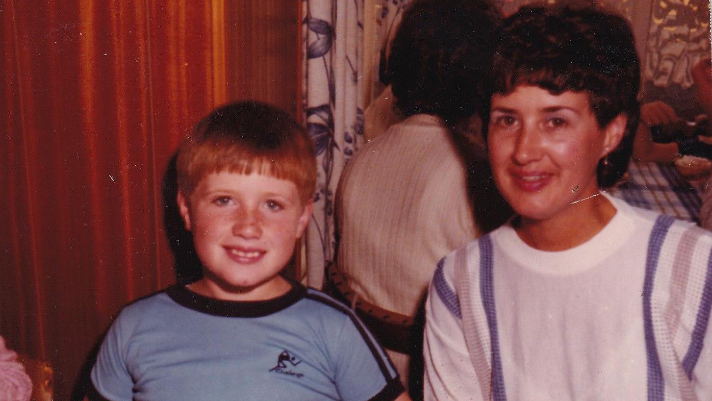 Photograph of a young Dale Morgan (left) pictured with his mother Judith Rhead (right). Judith has brown short hair and wears a white and purple sweater, while Dale wears a blue and black t-shirt and has short ginger hair and blue eyes. 