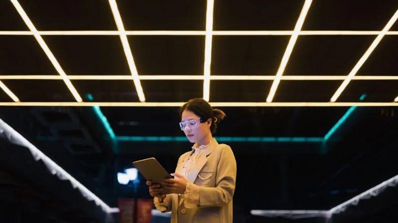 Una mujer con gafas transparentes y una tableta en la mano, en un salón oscuro con varias lámparas de luz fluorescente en el techo, las cuales le dan una sensación futurista a la foto. 