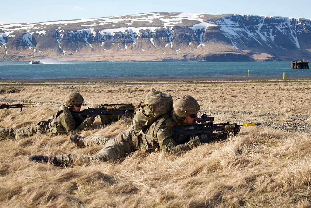 Dos soldados estadounidenses cuerpo a tierra apuntan con sus armas en una pradera. El situado en posición más atrasada parece una mujer. Al fondo hay una masa de agua y montañas moteadas de nieve. 