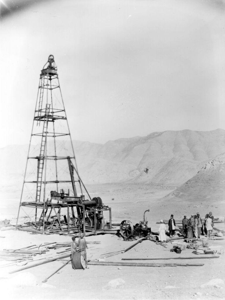 Una foto en blanco y negro muestra la construcción de una primitiva torre de extracción de petróleo en una región desértica con unas diez personas alrededor