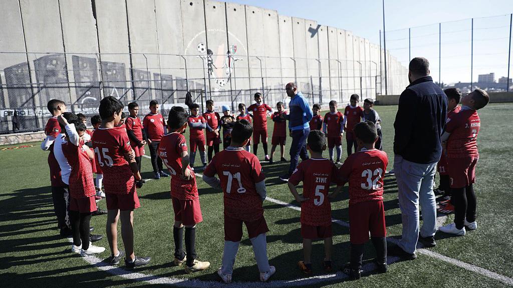 Un grupo de niños en uniforme de fútbol está parado en la mitad del campo, en un círculo, mientras escuchan al entrenador que les habla. 