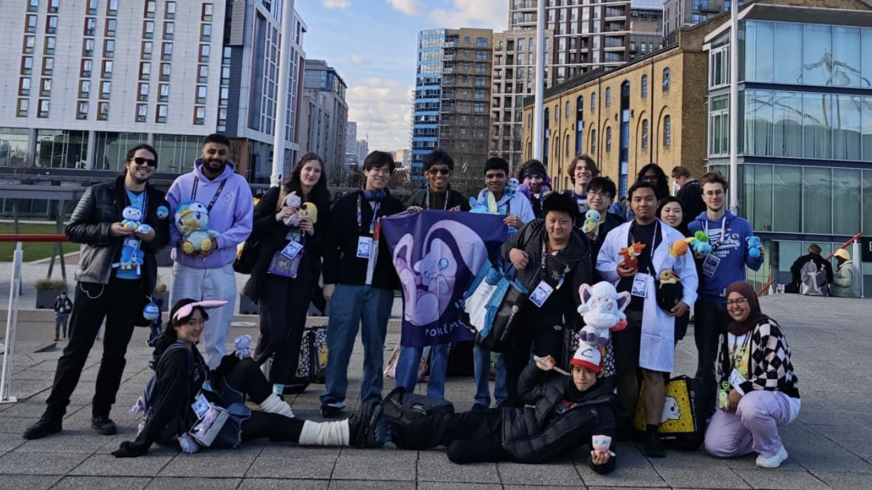 صورة تضم 17 عضواً من جمعية بوكيمون بجامعة لندن، يقفون في الهواء الطلق أمام مبانٍ شاهقة في المدينة. يحمل كل منهم دمية بوكيمون أو قطعة من البضائع التي تشير إلى شخصيتهم المفضلة.