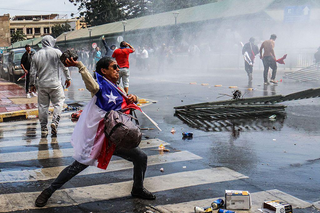 Un manifestante con la bandera chilena en el pecho arroja una piedra en una protesta.
