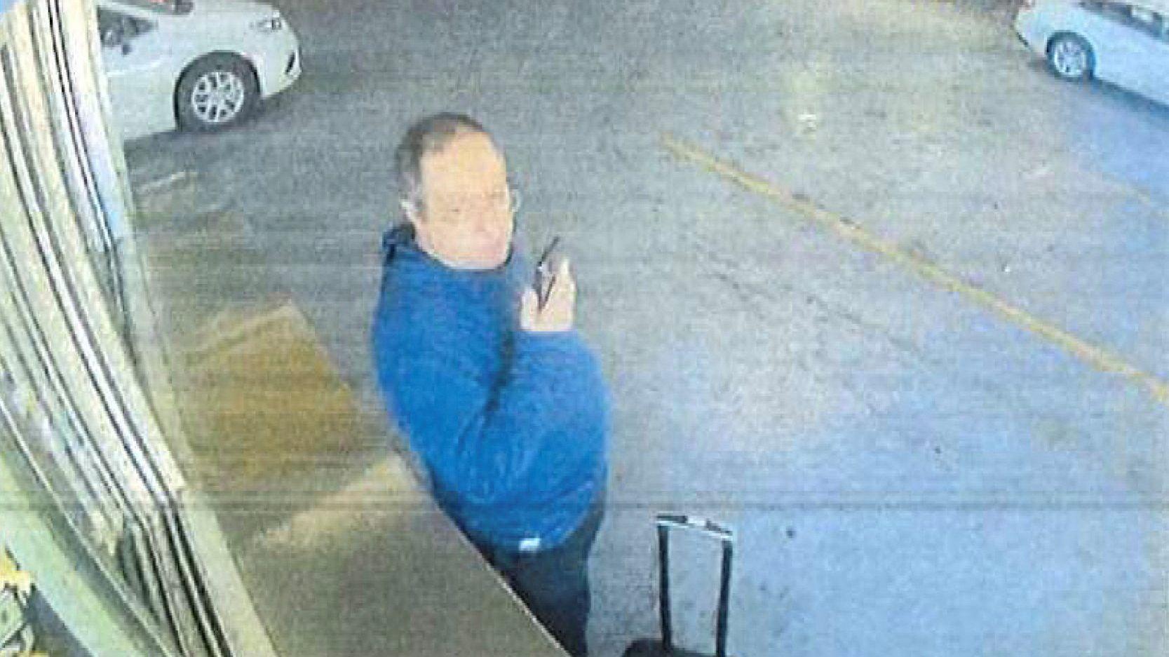 Uma pessoa segurando um telefone celular e vestindo um moletom azul aparece ao lado de uma mala com rodinhas, em um estacionamento subterrâneo, com carros ao fundo. 