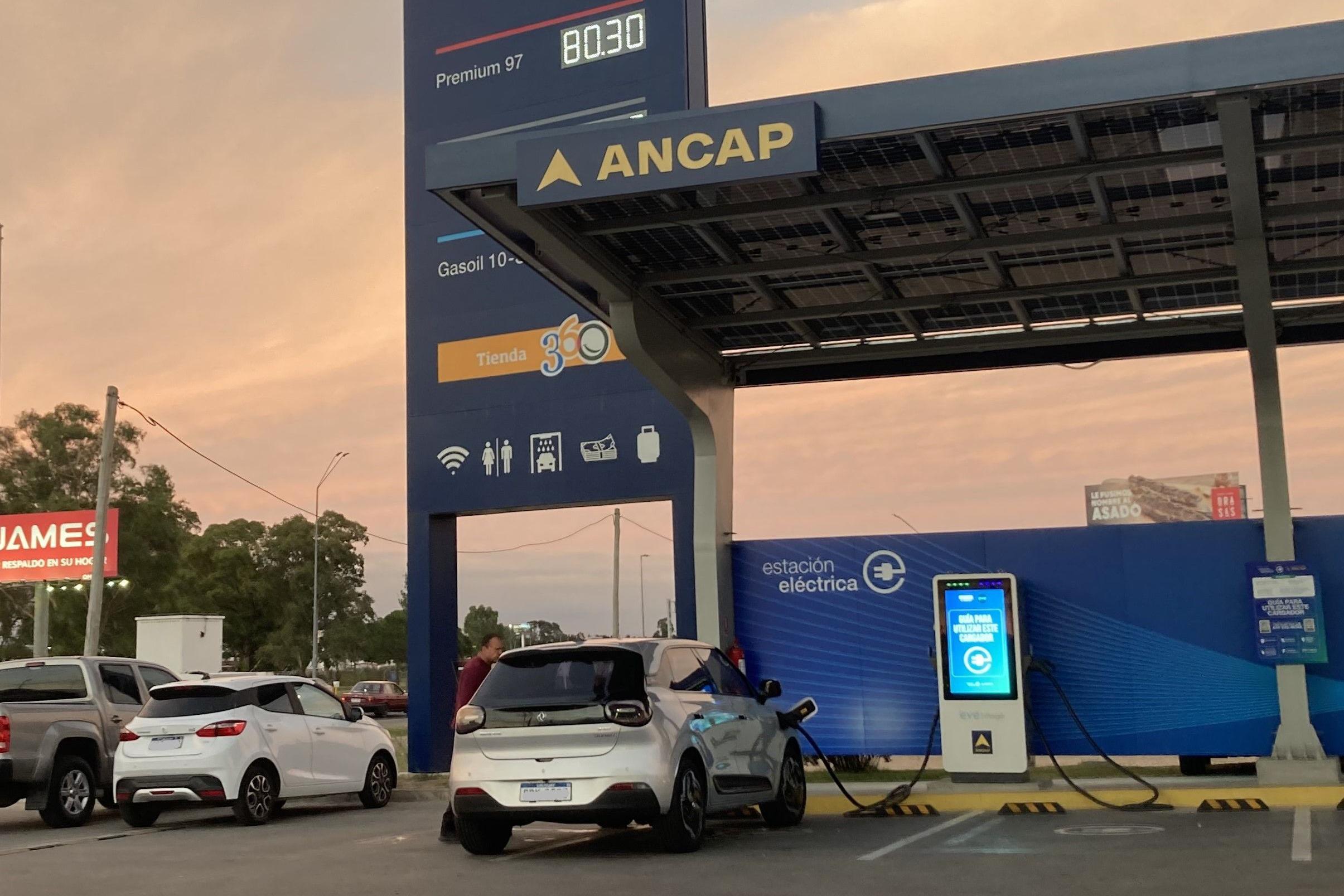 Carro el&eacute;trico carrega sua bateria em um posto com os pre&ccedil;os da gasolina no cartaz