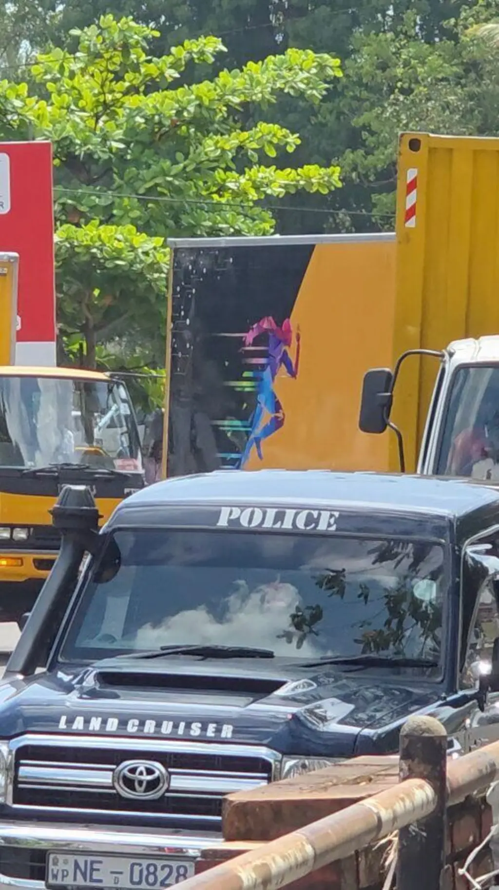පොලිස් ජීප් රථයක් ඉදිරියෙන් ගමන් කරන අතර කහ පැහැති ලොරි තුනක් පිටුපසින් පෙනේ.