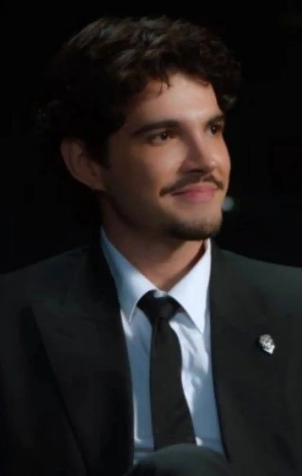 Jão, de cabelos escuros e bigode, veste terno preto, camisa branca e gravata preta, enquanto está sentado em uma poltrona. Ele sorri discretamente e olha para fora do enquadramento, em um ambiente escuro iluminado por luz direcionada. Copyright
