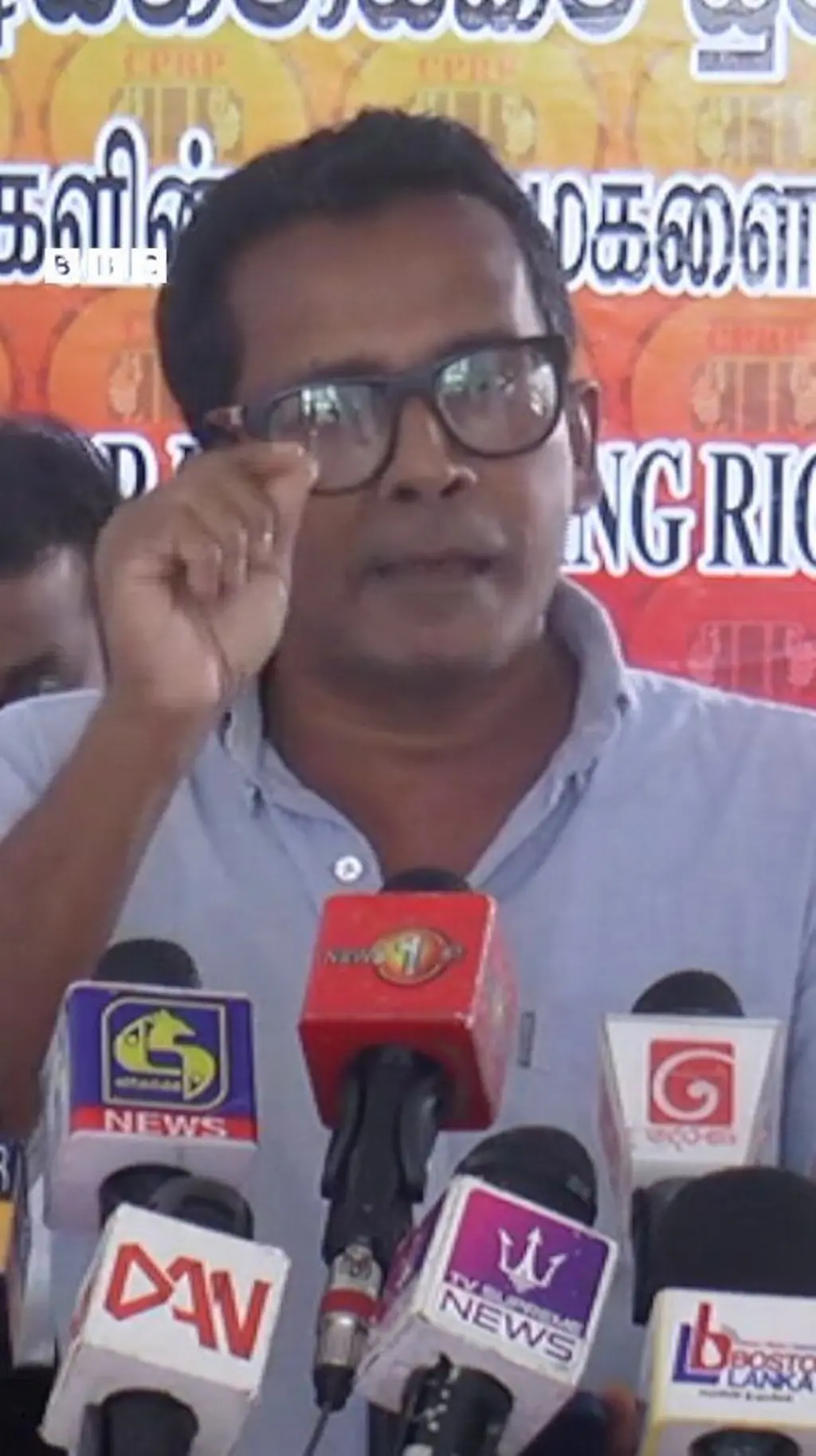 කණ්නාඩි දැමූ පුද්ගලයෙකු මාධ්‍ය හමුවක කතා කරනු පෙනේ.