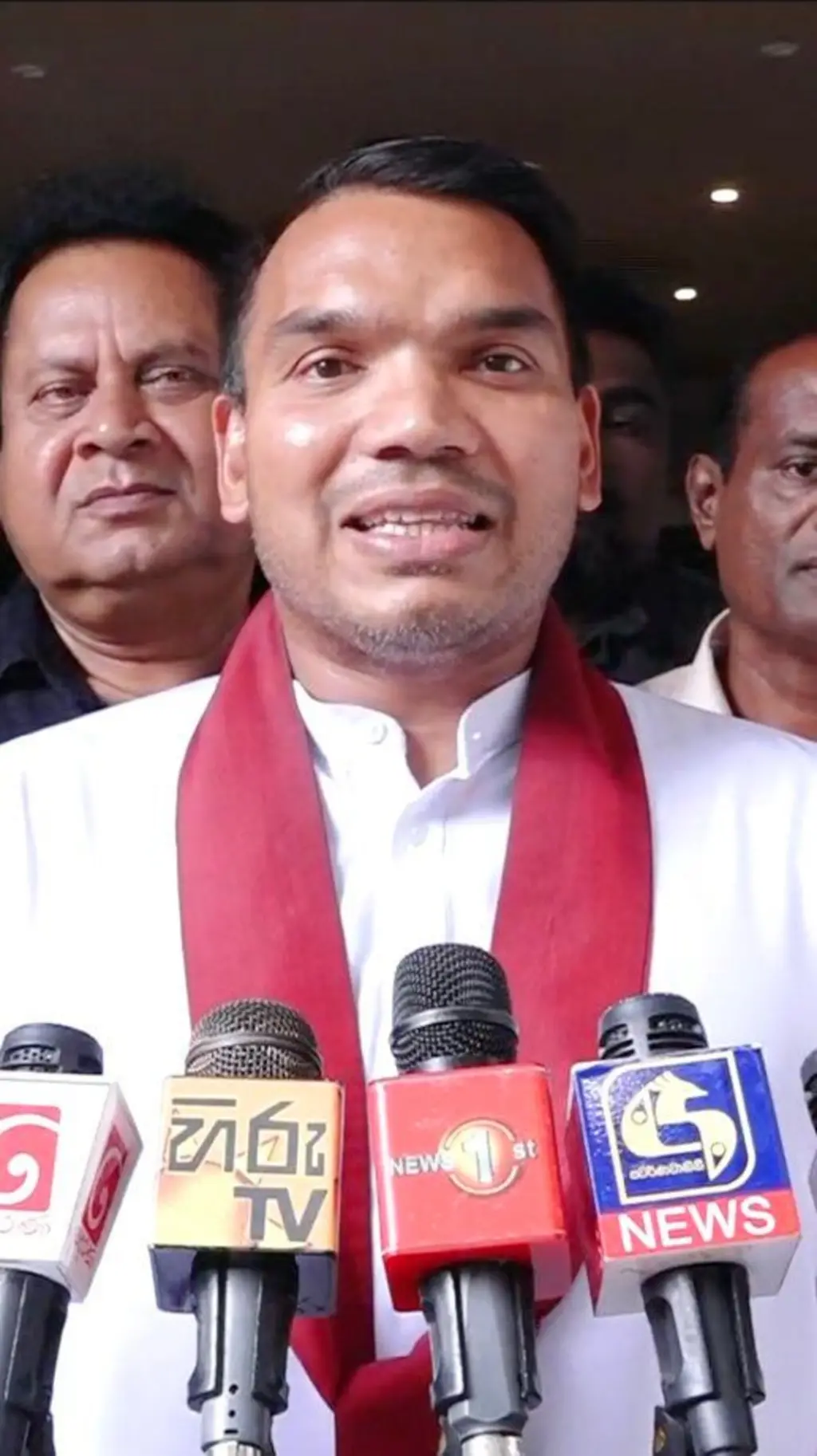 ජාතික වැඳුම ඇඳ, කුරහන් සාටකය පළඳි අයෙකු මක්‍රෆෝන හතරක් ඉදිරිපිට සිටී.