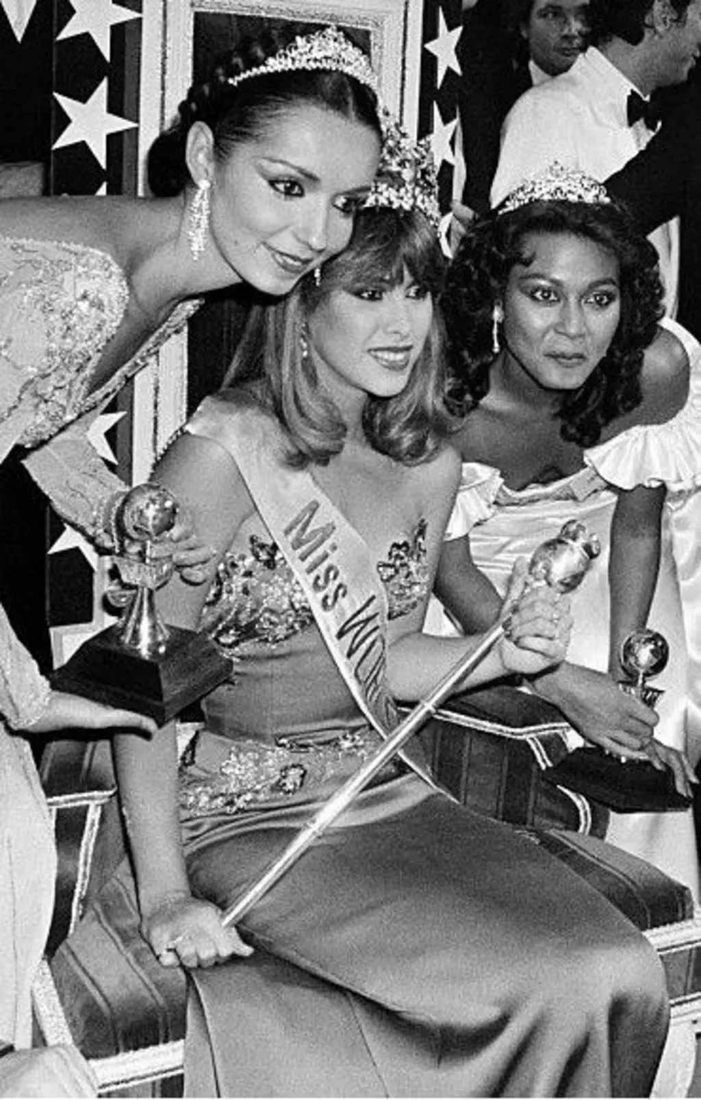 Mulher sentada em uma cadeira com uma coroa. Há várias pessoas ao redor