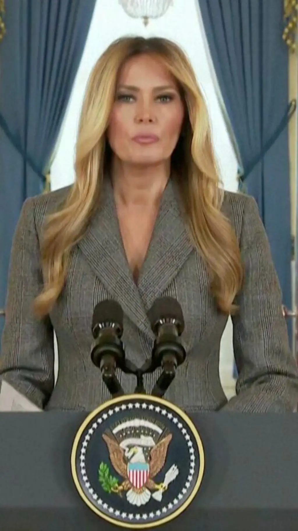 අළු පැහැති කබායකින් සැරසුණු මෙලනියා ට්‍රම්ප් මක්‍රෆෝනයක් ඉදිරිපිට සිටිනවා.