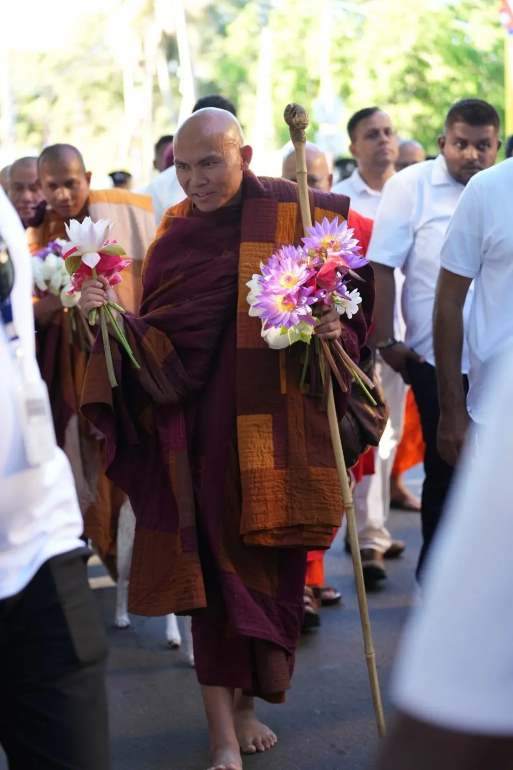 ස්වාමීන් වහන්සේ නමක් අත්දෙකෙහි නෙලුම් මල් සහ සැරයටියක් ගෙන පිරිසක් අතරින් ගමන් කරයි. 