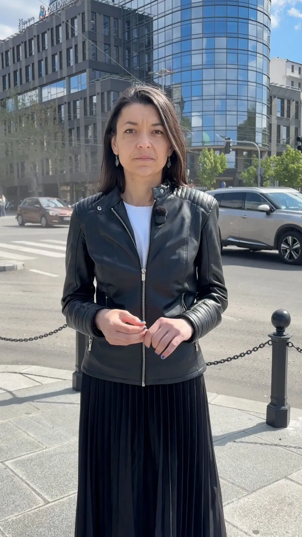 Novinarka stoji na trotoaru kod jedne od prometnijih raskrisnica u Beogradu. Iza nje se vide automobili koji prolaze.