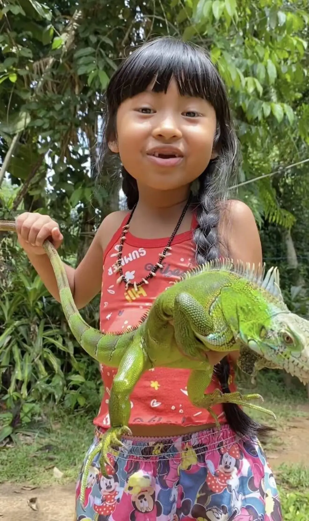 Fotografia colorida mostra a influencer Yandra Mawé, uma criança indígena, segurando uma iguana verde. Yandra sorri e tem uma trança no cabelo. O cenário ao fundo é de árvores e chão de terra.