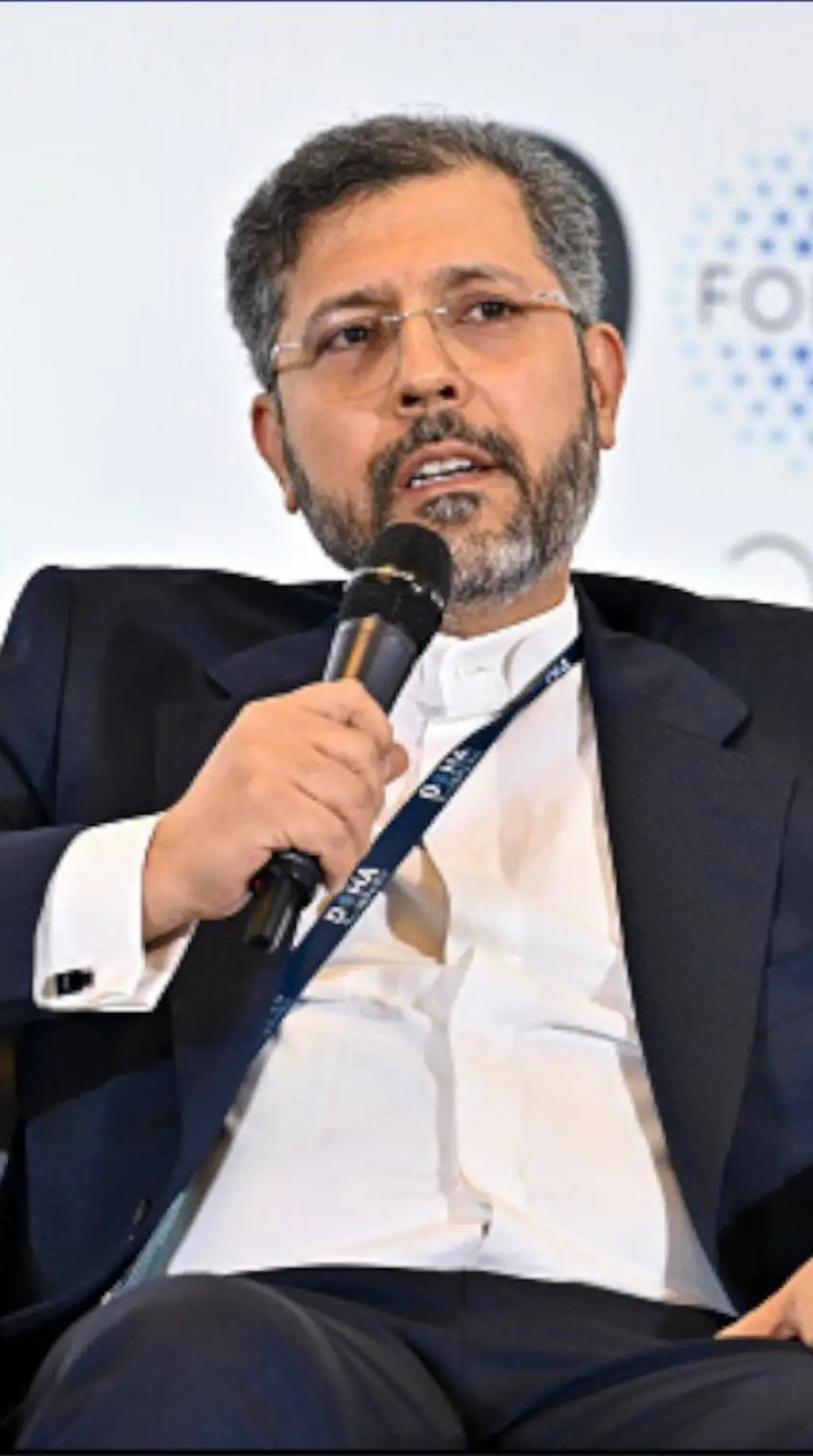 සුදු කමිසයකට උඩින් කළු කොට එකක් ඇඳ, කණ්නාඩි පැළඳ සිටින අයෙකු මක්රෆෝනයක් අතින්ගෙන සිටිනවා.