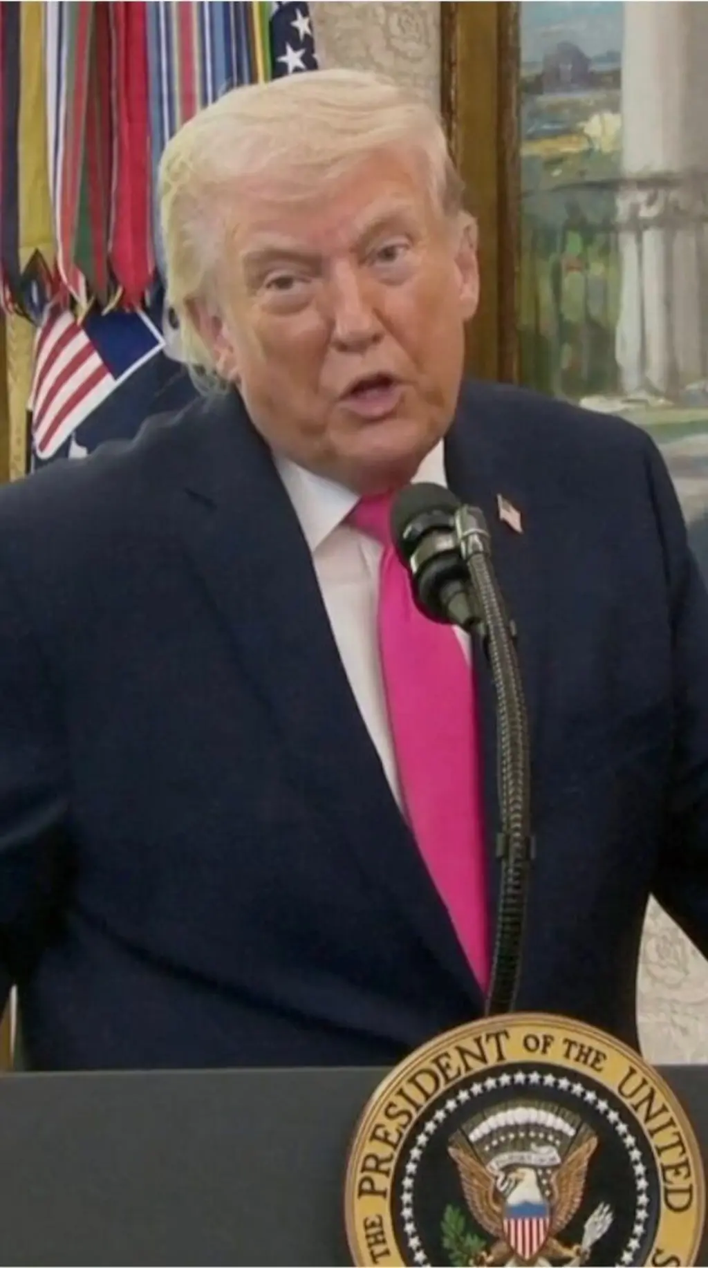 කළු කෝට් එකක්, සුදු පැහැති කමිසයක් සහ රෝස පාට ටයි එකක් පැළඳ සිටින ඇමෙරිකානු ජනාධිපති ඩොනල්ඩ් ට්රම්ප් මයික්රපෝනයක් ඉදිරිපිට කතා කරන ආකාරය දැක්වේ.