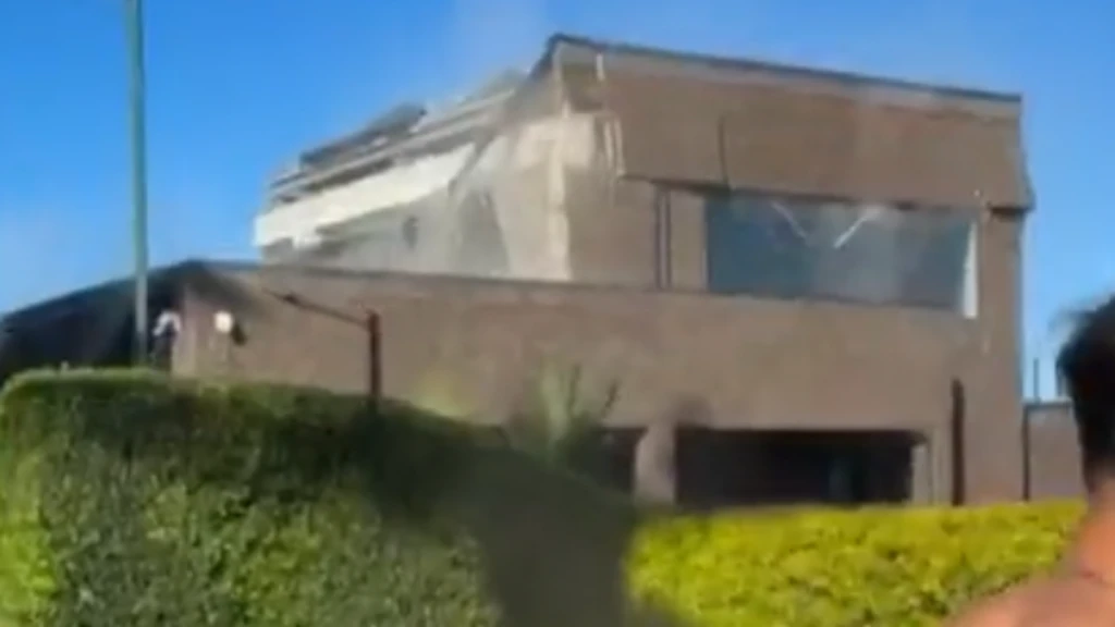 A imagem mostra um edifício moderno de fachada marrom, parcialmente encoberto por uma cerca viva, sob um céu azul. Ao fundo, há uma estrutura de aparência industrial ou institucional.