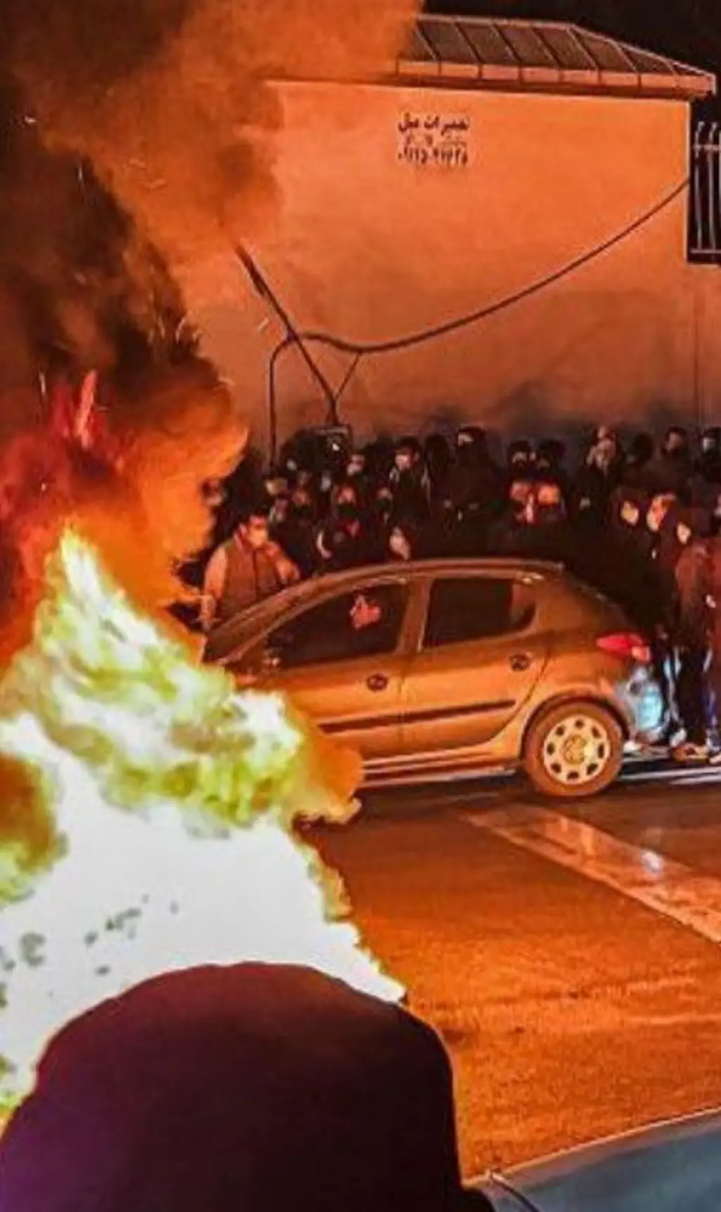 Fogo na rua. Ao redor há várias pessoas e um carro. 