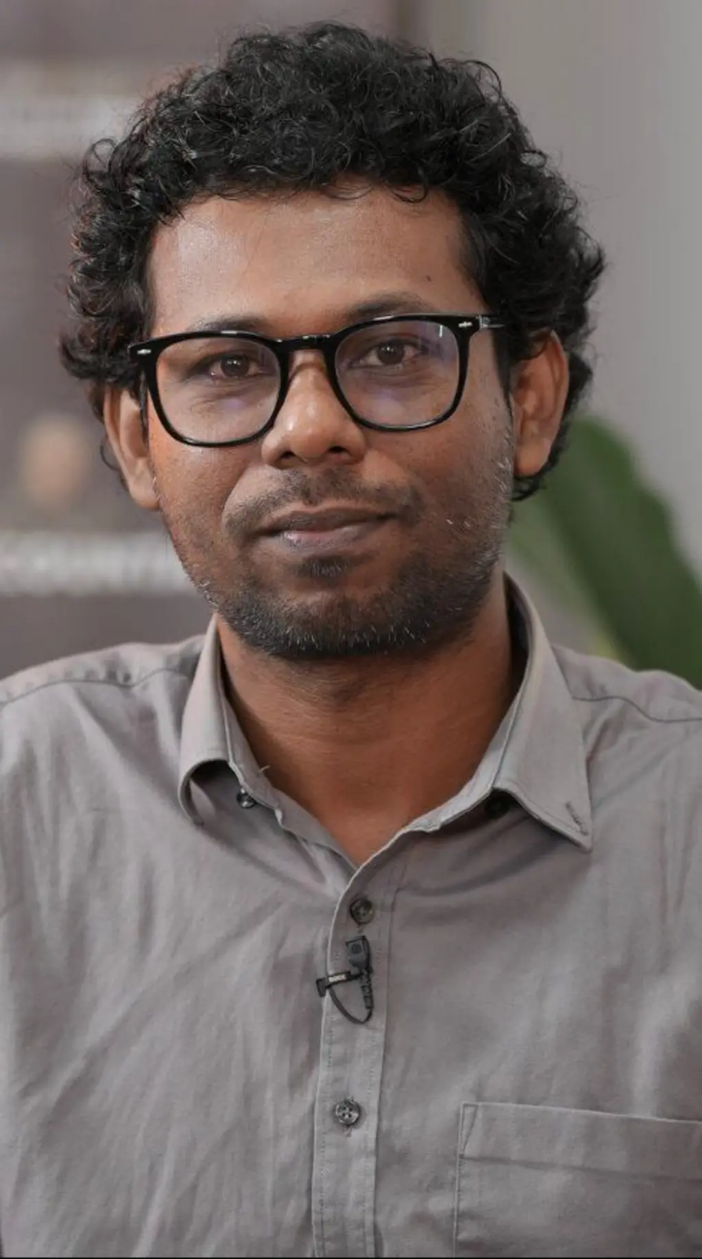 ක්‍රේල් කොණ්ඩයක් සහිත, කණ්නාඩි පැළඳ සිටින අයෙකු අළු පාට කමිසයක් ඇඳ සිටී.