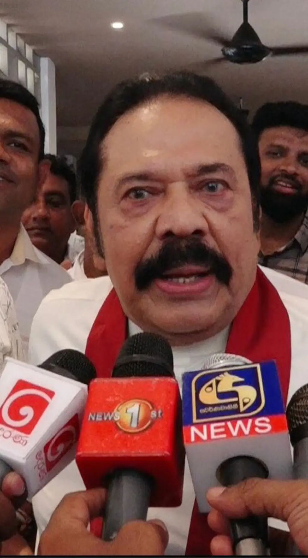 ජාතික ඇඳුම සහ කුරහන්පාට සාටකය පැළඳි මහින්ද රාජපක්ෂ මයික්‍රපෝන තුනක් ඉදිරිපිට කතා කරන ආකාරය පෙනේ.