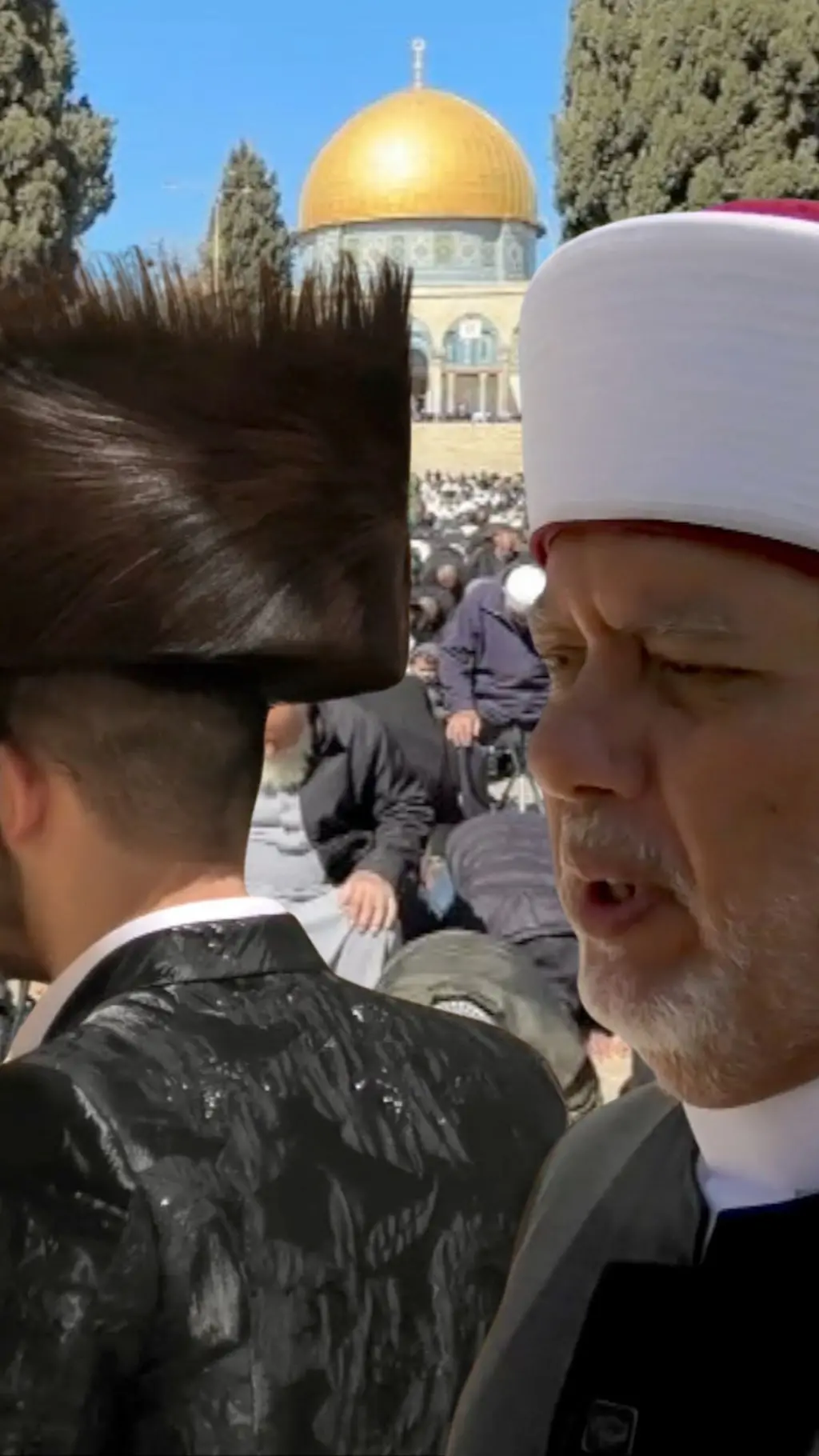 Rasnda Qudduslik mulla, bitta yahudiy bosh kiyimini kiygan ziyoratchi va ularning orqasida Baytul Muqaddas gumbazi ko'rinadi