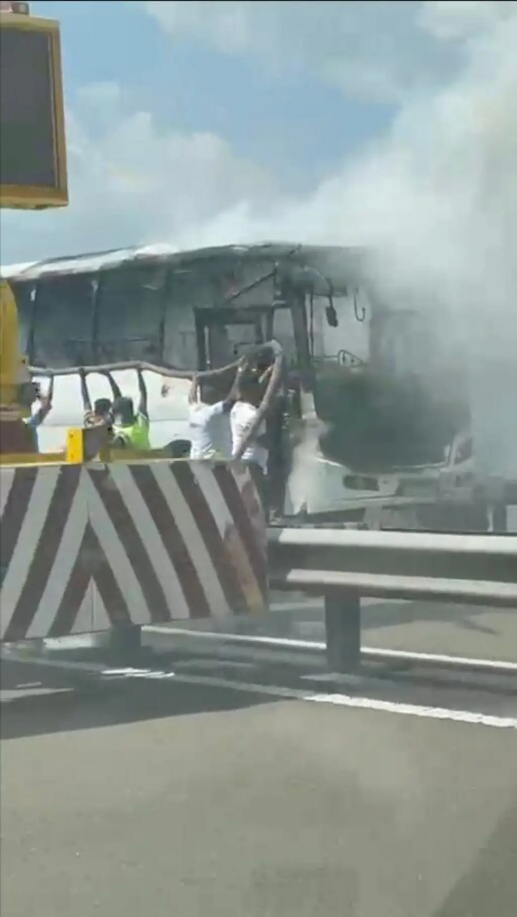 මාර්ගයක සුදු පැහැති බස් රථයක් ගිනි ගන්නා අතර මිනිසුන් වතුර බටයක් ගෙන එය නිවීමට උත්සාහ කරන අයුරු පෙනේ.