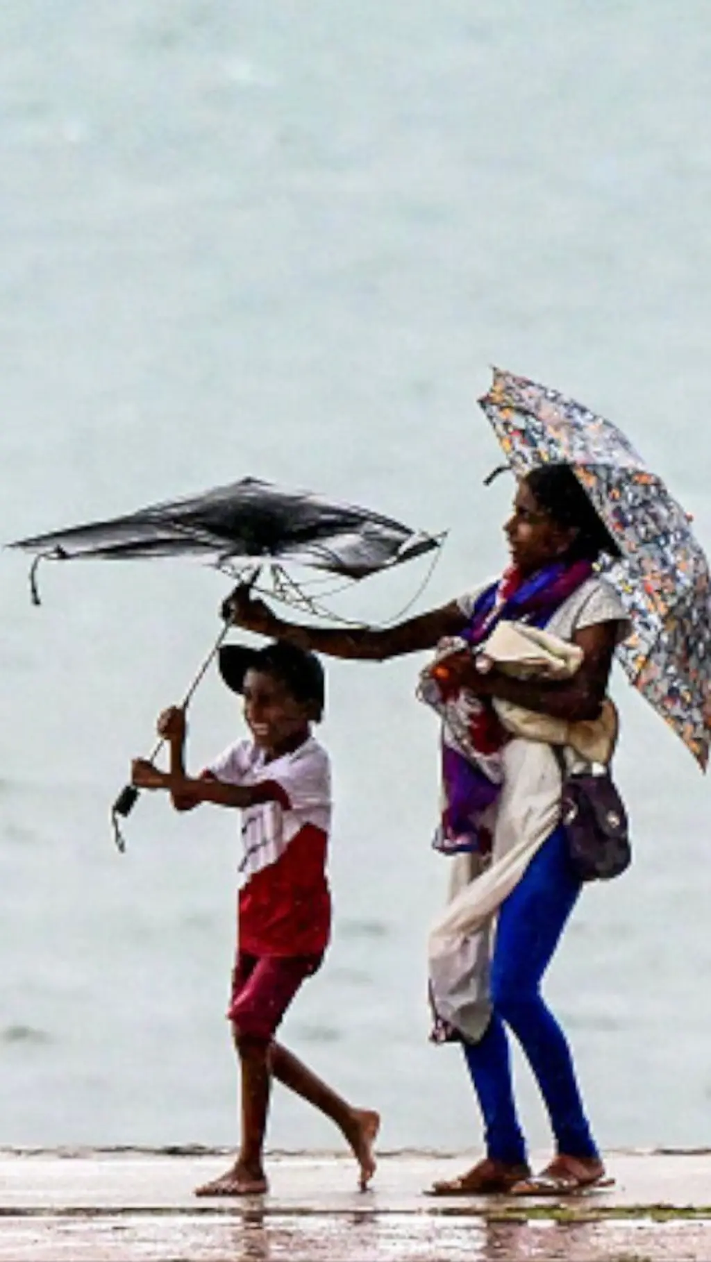 කාන්තාවක් සහ කුඩා පිරී දරුවෙක් වැස්ස අවස්ථාවක මුහුදක් අසලින් ගමන් කරන අතර දරුවාගේ කුඩය සුළඟට ගසා ගෙන යන අයුරු පෙනේ.