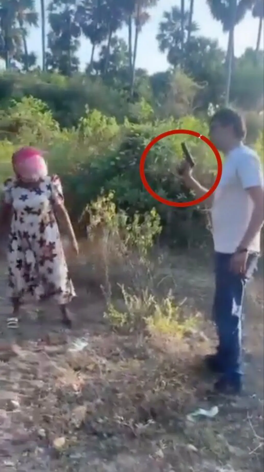 සුදු ටී ෂර්ට් එකක් සහ කළු දිග කලිසමක් ඇඳ සිටින අයෙකු හෙල්මට් එකක් දාගෙන ඉන්න කන්ත්වකට පිස්තෝලයක් පෙන්වන අයුරු පෙනේ.