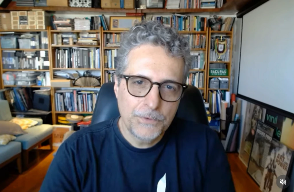 Kleber Mendonça Filho dando entrevista via chamada de vídeo. Ele está sentado e atrás dele há uma estante com livros