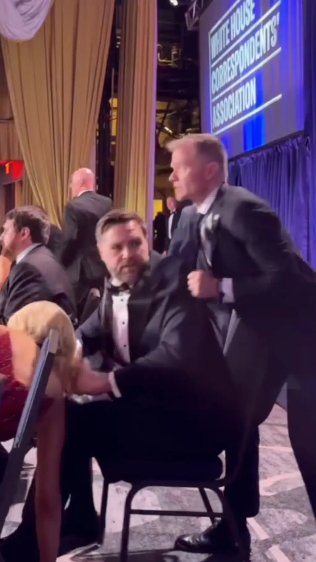 පුටුවක වාඩි වී සිටින කෝට එකක් ඇඳගත් පුද්ගලයෙකුව තවත් පුද්ගලයෙකු විසින් එසවීමට උත්සාහ කරන ආකාරය පෙනේ