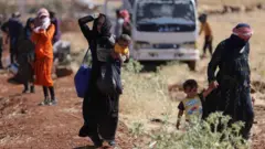 Mujeres beduinas con niños caminan por un sendero de tierra en el sur de Siria. Una lleva de la mano a un niño pequeño y otra lleva a un bebé en brazos.