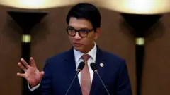 Umugabo wambaye indorerwamo ziriho umurongo (umuguno) w'umukara hamwe na kositimu y'ubururu na karuvati itukura, arimo kuvugira mu ndangururamajwi ebyiri, azamuye ikiganza cye mu gihe arimo kuvuga. 

