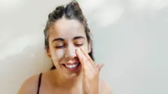 Retrato en primer plano de una joven aplicándose crema en las mejillas y la frente.